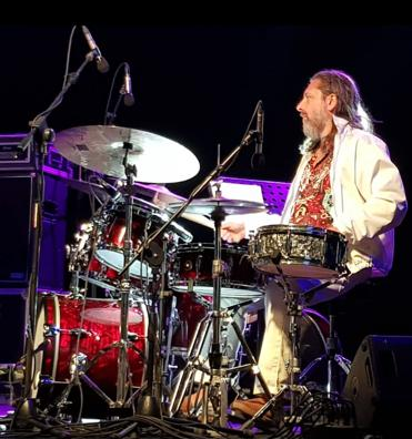 Batterista sul palco che suona una batteria rossa. Indossa una giacca bianca, ha i capelli lunghi e la barba.