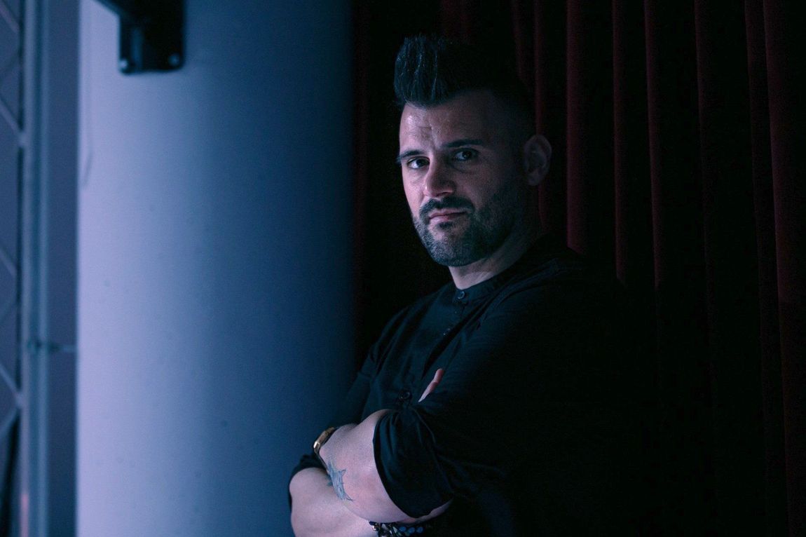 Un uomo con la cresta, braccia incrociate, guarda la telecamera. Si trova in una stanza scarsamente illuminata, con pareti blu e tende rosse.