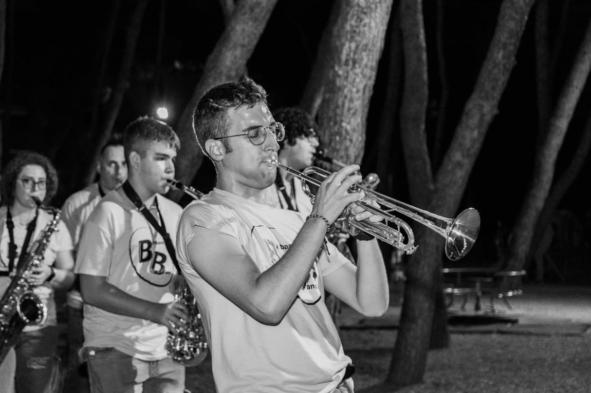 La banda suona all'aperto di notte; il trombettista è in primo piano, sullo sfondo ci sono degli alberi.