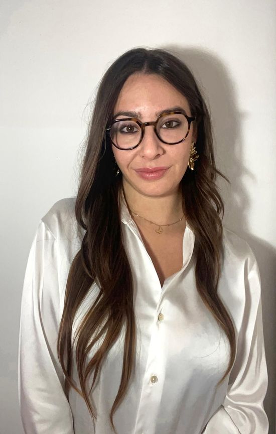 Una donna con gli occhiali e una camicia bianca abbottonata sorride alla telecamera.