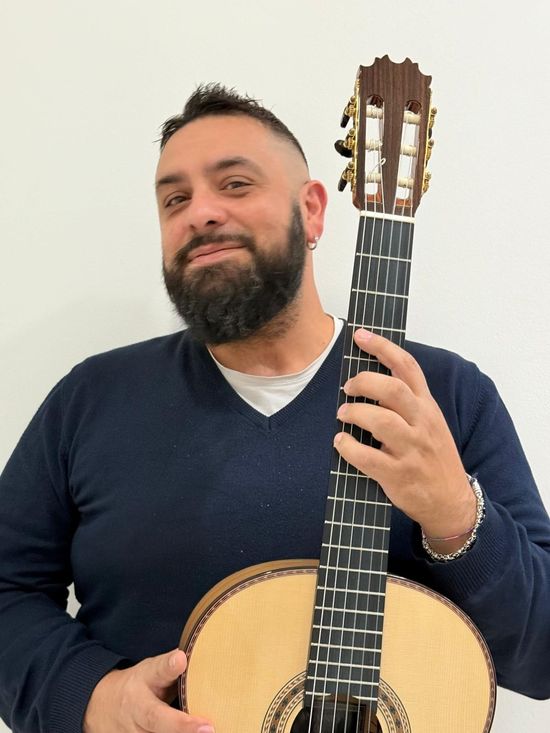 Uomo che tiene in mano una chitarra, sorridente. Indossa un maglione blu e ha la barba.