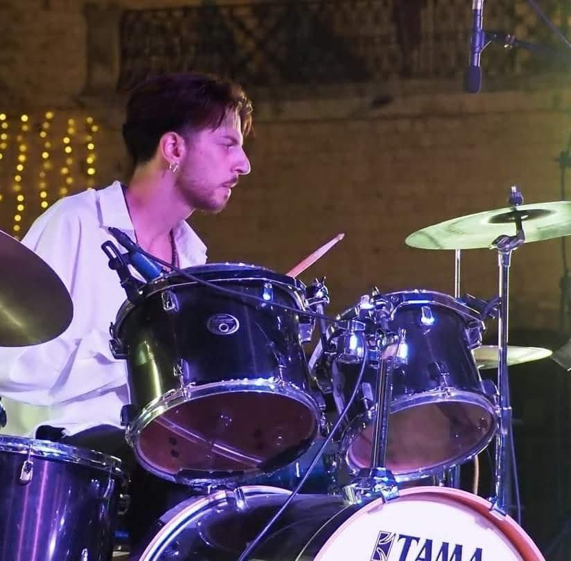 Un batterista suona una batteria Tama nera all'aperto. Indossa una camicia bianca e ha un'espressione concentrata.