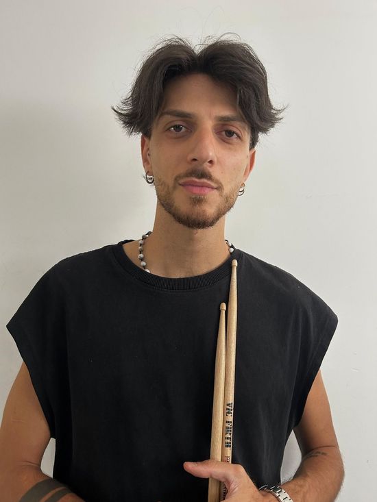 Uomo che tiene in mano delle bacchette, indossa una maglietta nera senza maniche e guarda la telecamera.