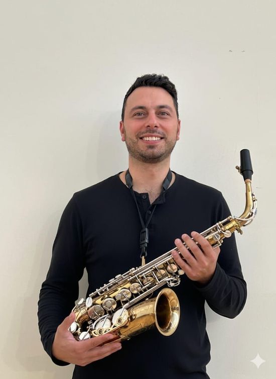 Uomo sorridente che tiene in mano un sassofono. Indossa abiti neri e sfondo bianco.