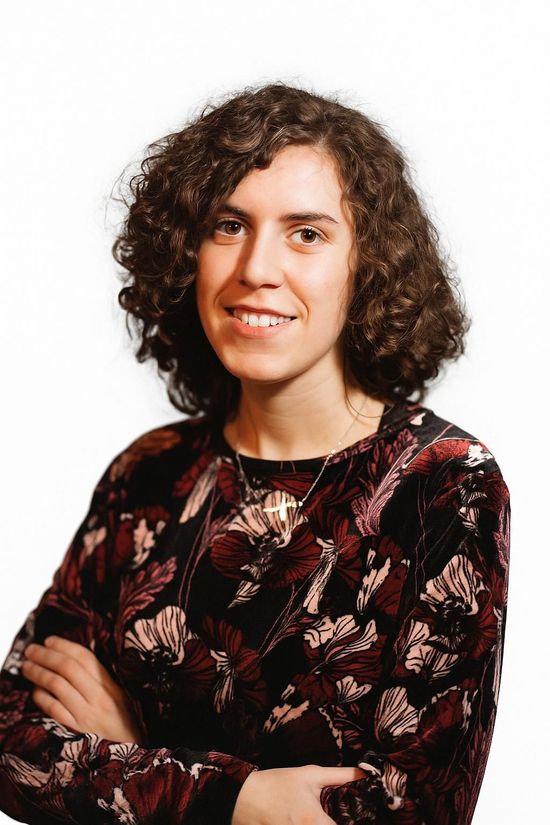 Donna con capelli castani ricci, braccia incrociate, sorridente. Indossa una camicia bordeaux e nera a fantasia.