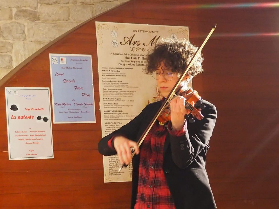 Persona che suona il violino, in un ambiente chiuso, con occhiali e una camicia a quadri rossa, con dei poster appesi alla parete dietro di sé.