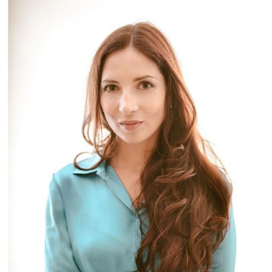 Donna con lunghi capelli castani, che indossa una camicia turchese abbottonata, che guarda la telecamera.