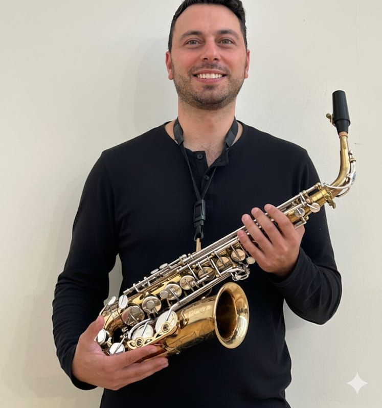 Uomo che tiene in mano un sassofono, sorridente, indossa una camicia nera su uno sfondo chiaro.