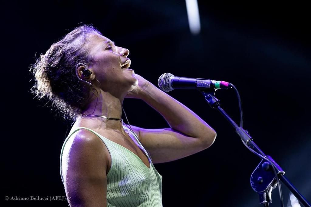 Donna che canta al microfono sul palco, sorride con la testa reclinata all'indietro e indossa una maglietta verde chiaro.
