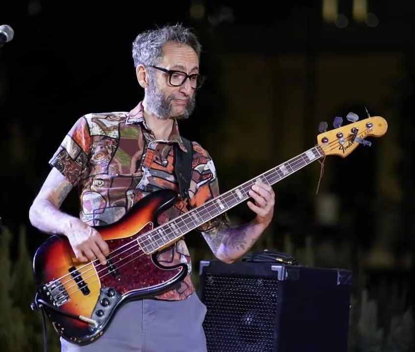 Uomo che suona il basso elettrico all'aperto di notte; indossa occhiali e una camicia a fantasia.