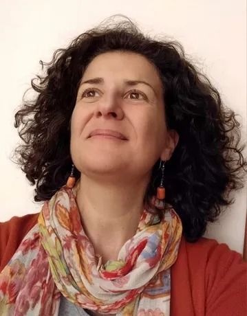 Donna con i capelli castani ricci, che guarda verso l'alto e sorride; indossa una sciarpa colorata e un cardigan arancione.