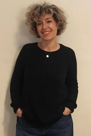 Donna sorridente con i capelli ricci, mani nelle tasche dei jeans, indossa un maglione nero.