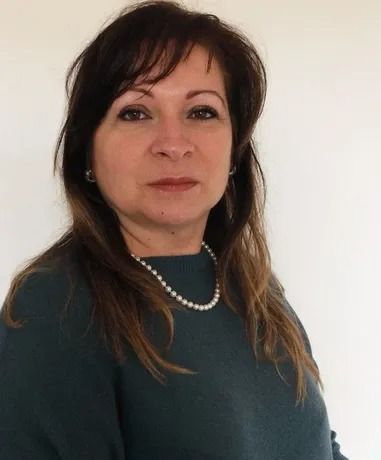 Donna con capelli castani, maglione verde e collana di perle, che guarda avanti, su sfondo bianco.