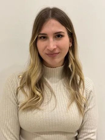 Donna con lunghi capelli biondi e ondulati, che indossa un dolcevita color crema, sorride leggermente, su uno sfondo bianco.