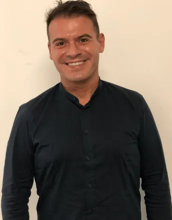 Un uomo con una camicia blu scuro sorride alla telecamera su uno sfondo bianco.