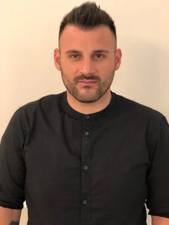 Uomo con capelli scuri, barba, che indossa una camicia nera, guarda la telecamera.