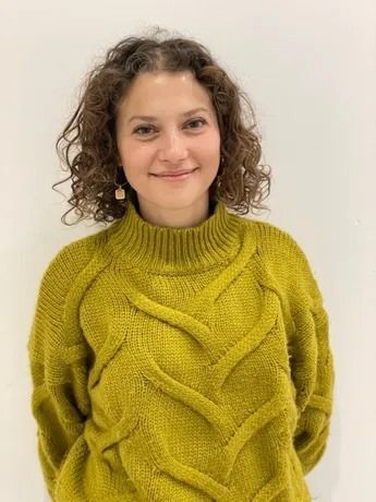 Donna con i capelli ricci, con un maglione verde lavorato a maglia, sorridente, su sfondo bianco.