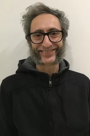 Uomo con occhiali e basette, sorridente, che indossa una giacca nera, su sfondo bianco.