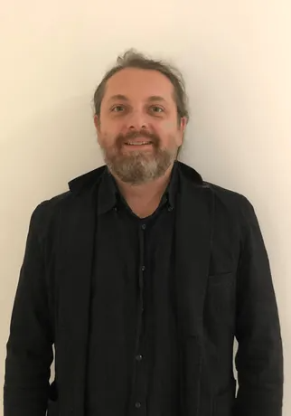 Uomo con la barba e una camicia nera che sorride, su sfondo bianco.