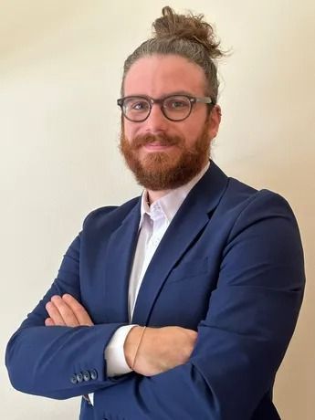 Uomo con barba e chignon, occhiali, abito blu, braccia incrociate, sorridente.