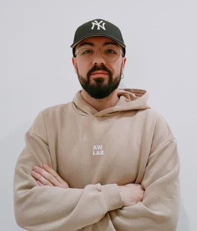 Uomo che indossa un berretto da baseball nero e una felpa con cappuccio marrone chiaro, con le braccia incrociate e rivolto in avanti.