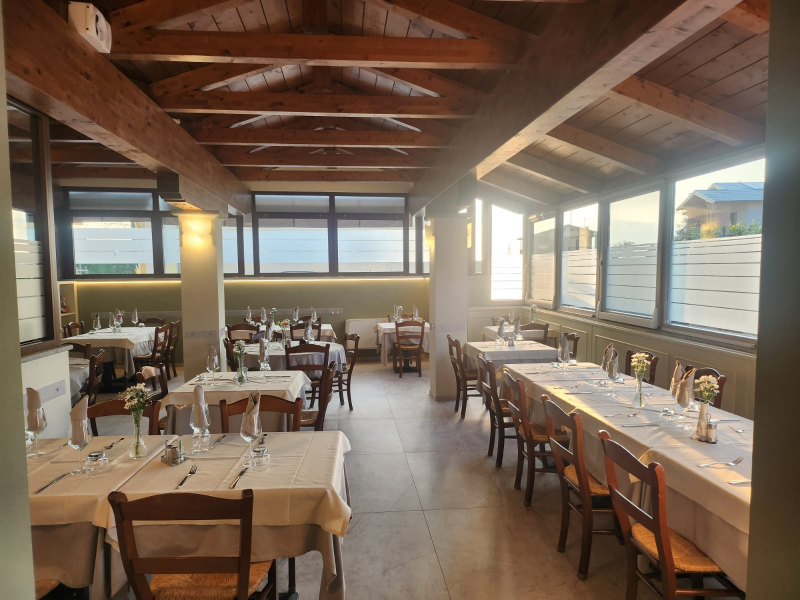 Luminosa sala da pranzo del ristorante con travi in ​​legno, lunghi tavoli apparecchiati e luce naturale che filtra dalle grandi finestre.