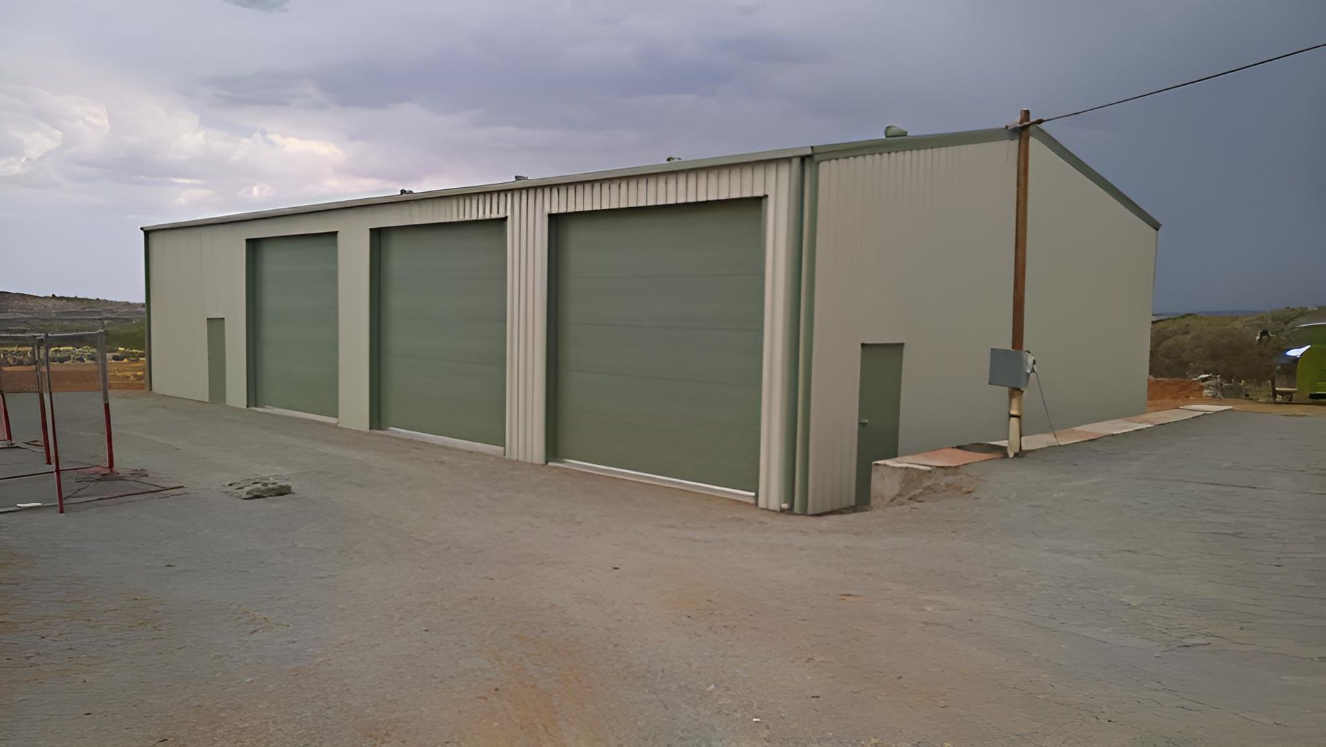 A Large Building With Green Doors Is Sitting On The Side Of A Dirt Road — Alice Sheds & Structures In Ciccone, NT
