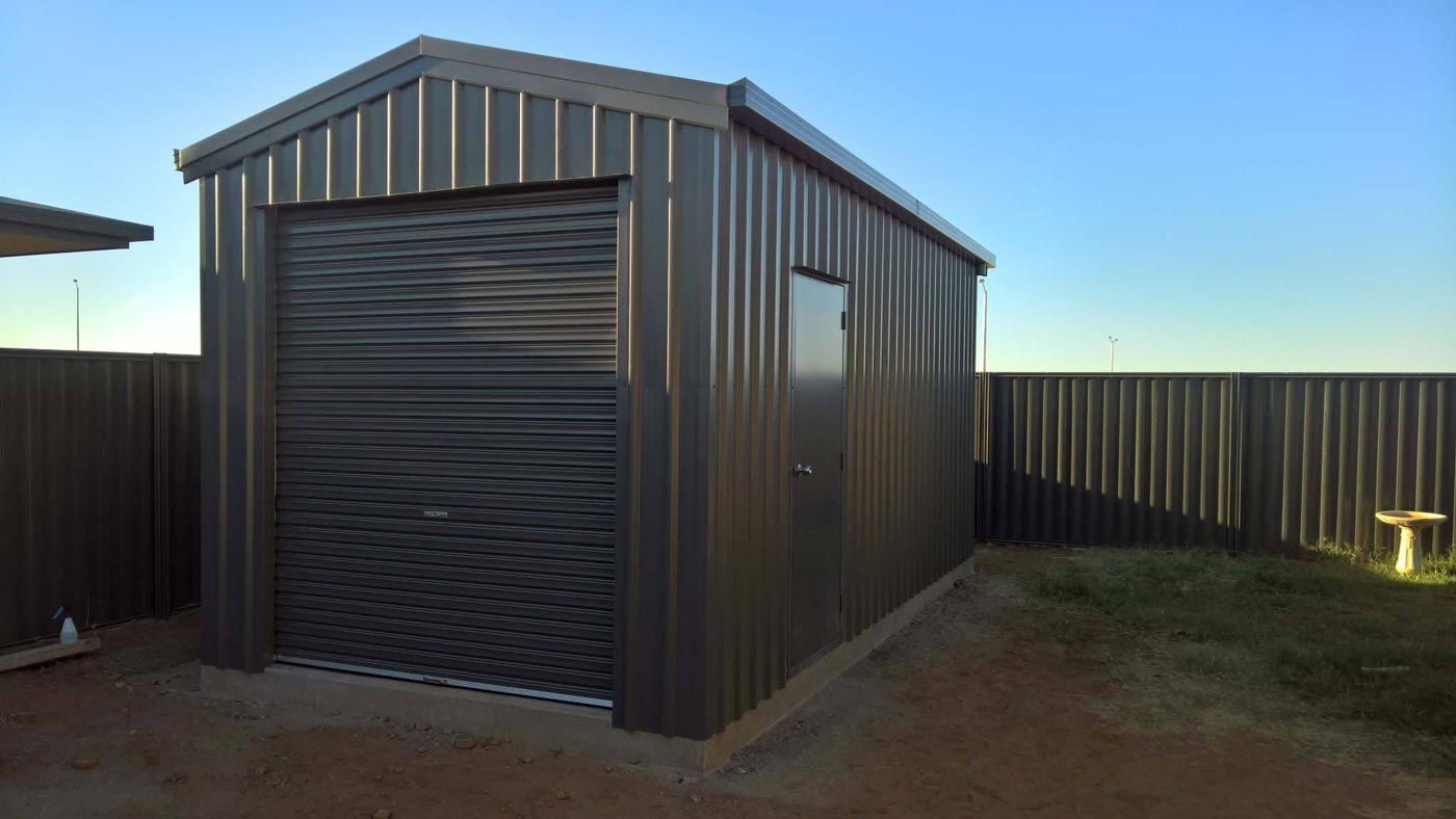 A Shed With A Garage Door And A Fence In The Background — Alice Sheds & Structures In Ciccone, NT