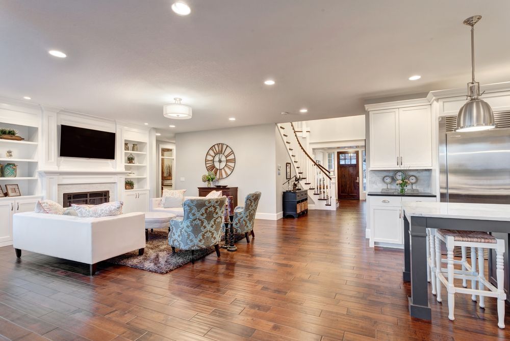 Bright open-concept living room and kitchen with white furniture, wood floors, and staircase.