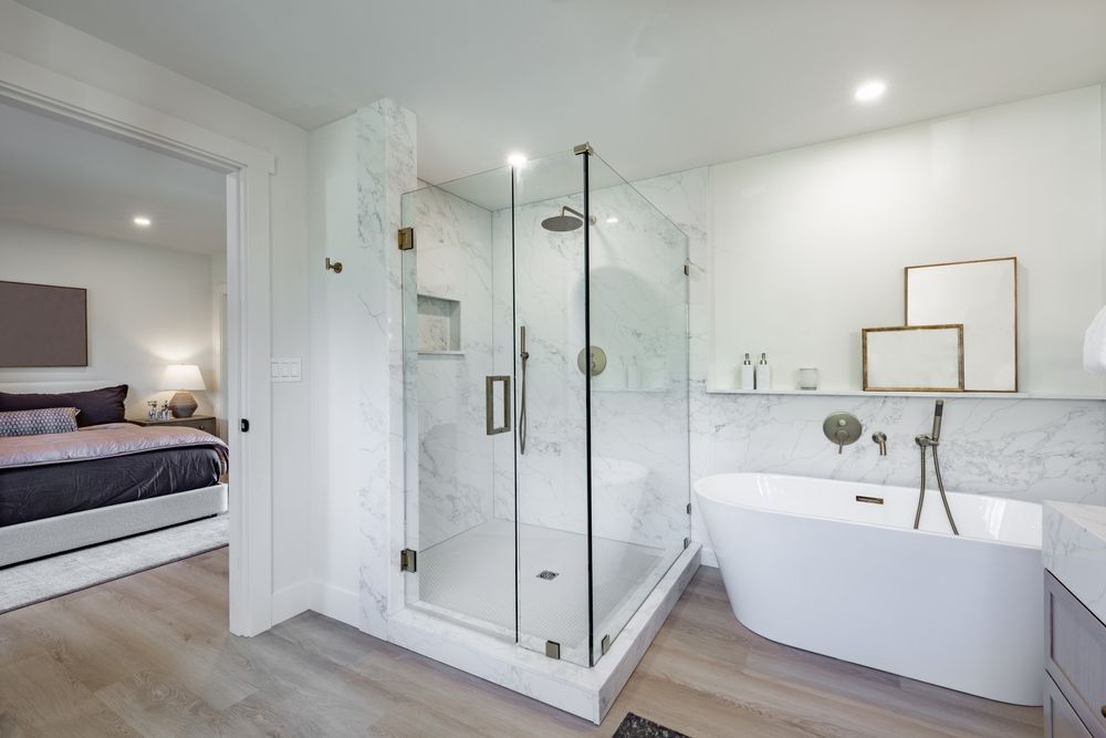 Modern white bathroom with glass shower, freestanding tub, and bedroom visible through open doorway