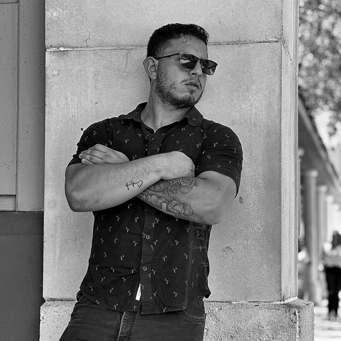 Man with crossed arms, sunglasses, leaning against a column outdoors.