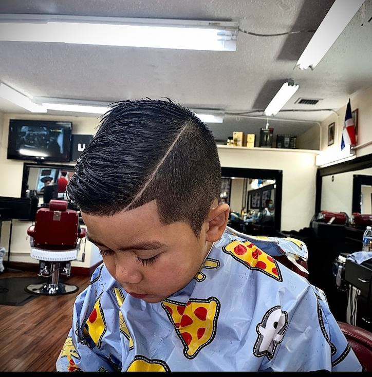 Child with a fade haircut in a barbershop, wearing a pizza-patterned cape.