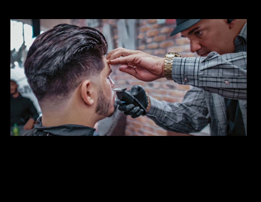 Barber trimming a man's beard with clippers in a barbershop. The man wears a black cape. The barber wears a hat.