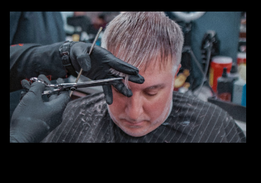 Barber cutting hair in a shop; hands in black gloves with scissors; client wearing a cape.