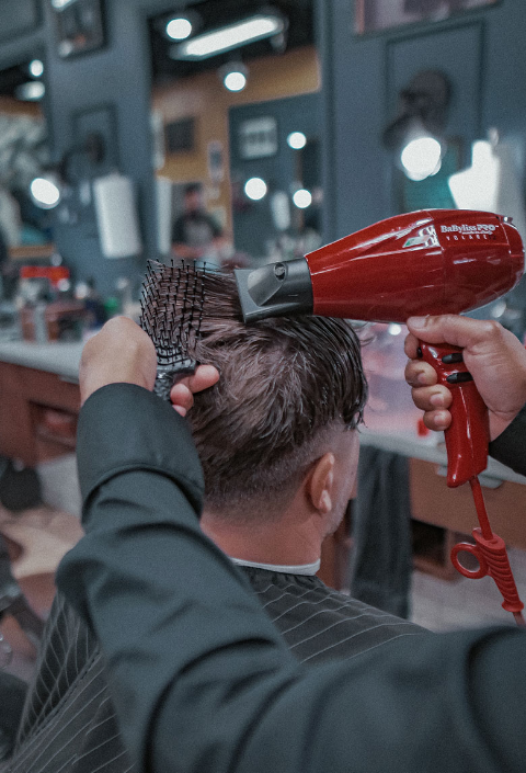 Barber using a red hairdryer and brush to style a client's hair in a barbershop.