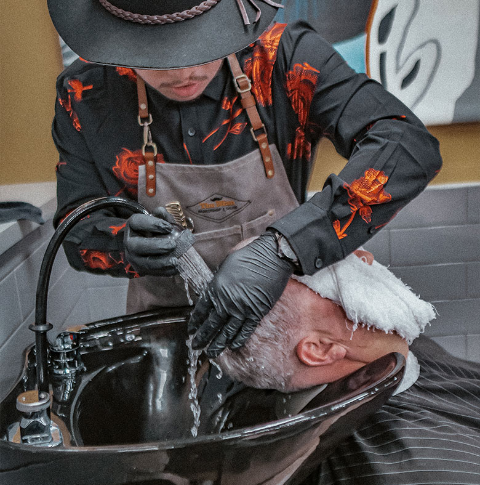 Barber washing customer's hair in a salon. The barber wears a hat, apron, and gloves.