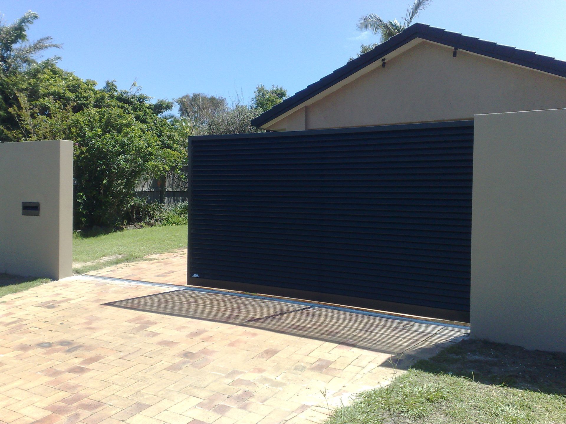 Dark Metal Sliding Gate Between Beige Walls, in Front of a House — Coastal Auto Doors & Gates In Palmwoods, QLD