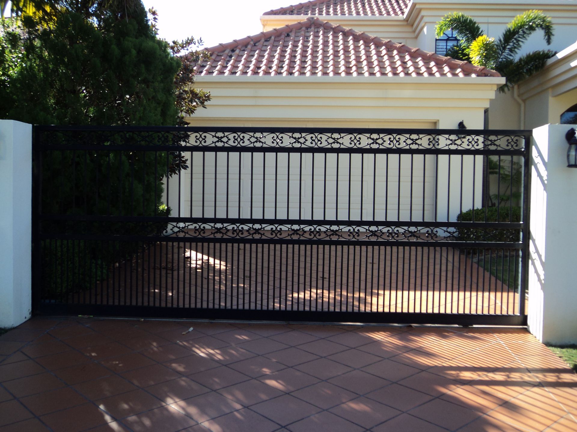 Black Metal Driveway Gate in Front of a Tan House With Red Tile Roof — Coastal Auto Doors & Gates In Palmwoods, QLD