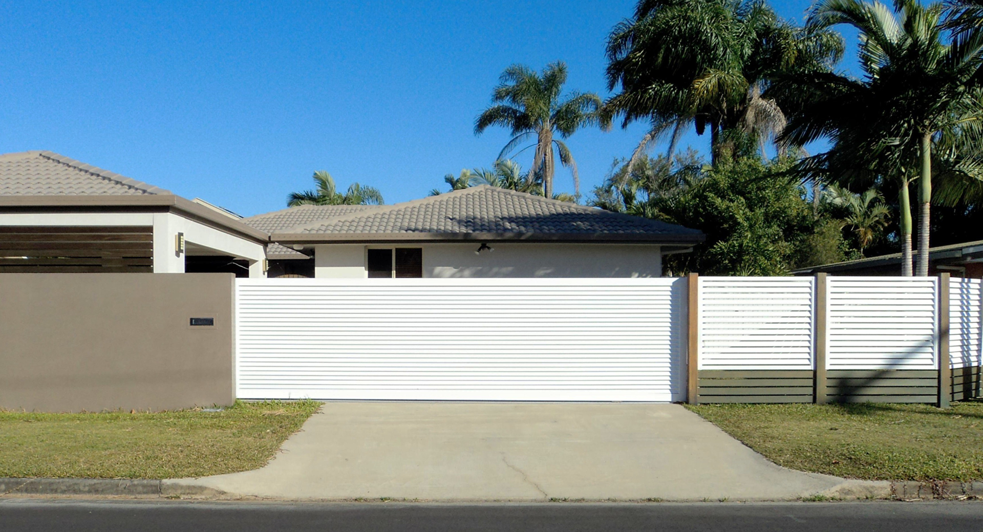White horizontal slat driveway gate and fence in front of a house, with blue sky and trees in background — Coastal Auto Doors & Gates In Caloundra, QLD