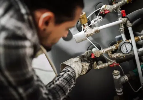 A person in a plaid shirt wearing work gloves adjusts valves on a gray mechanical heating system.