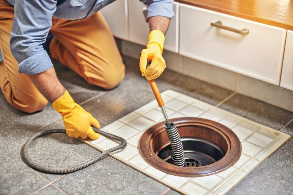 A person wearing yellow gloves uses a long brush to clean a drain in a tiled floor.