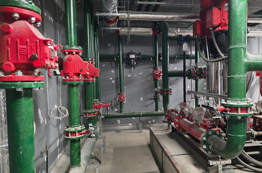 An industrial mechanical room with green piping, red valves, and pumps installed against grey walls.