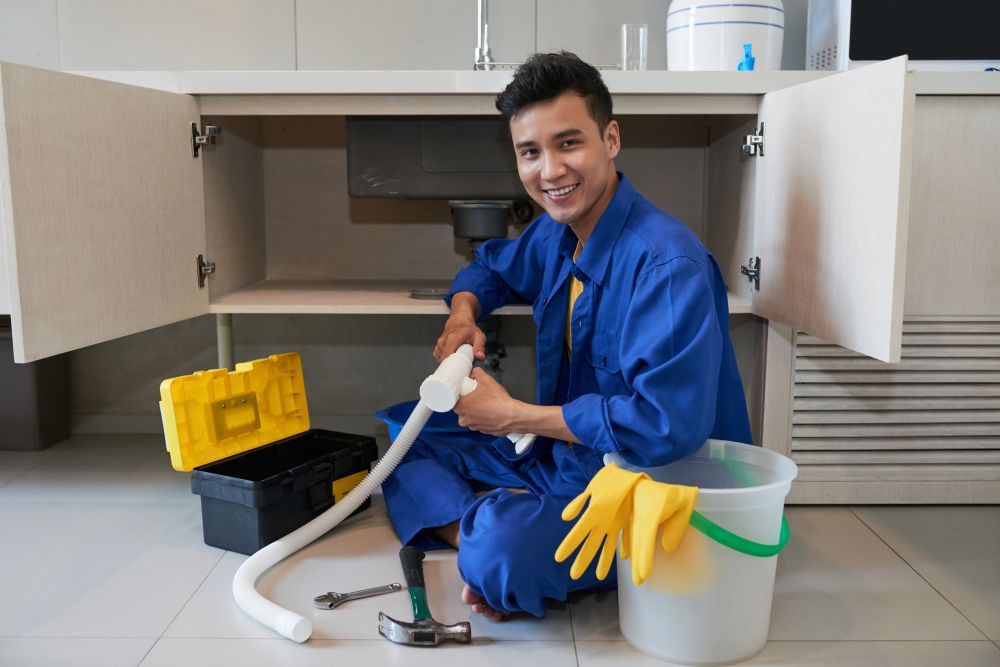 A smiling plumber in blue coveralls sits on the floor fixing a sink pipe, surrounded by a toolbox and cleaning supplies.
