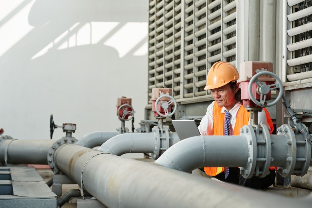 A person wearing a hard hat and high-visibility vest uses a laptop while inspecting industrial pipes near a cooling tower.