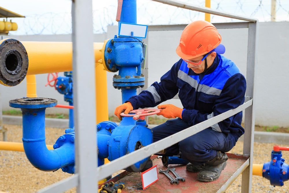 A worker in a hard hat and protective uniform uses tools on blue industrial valves at a gas facility.