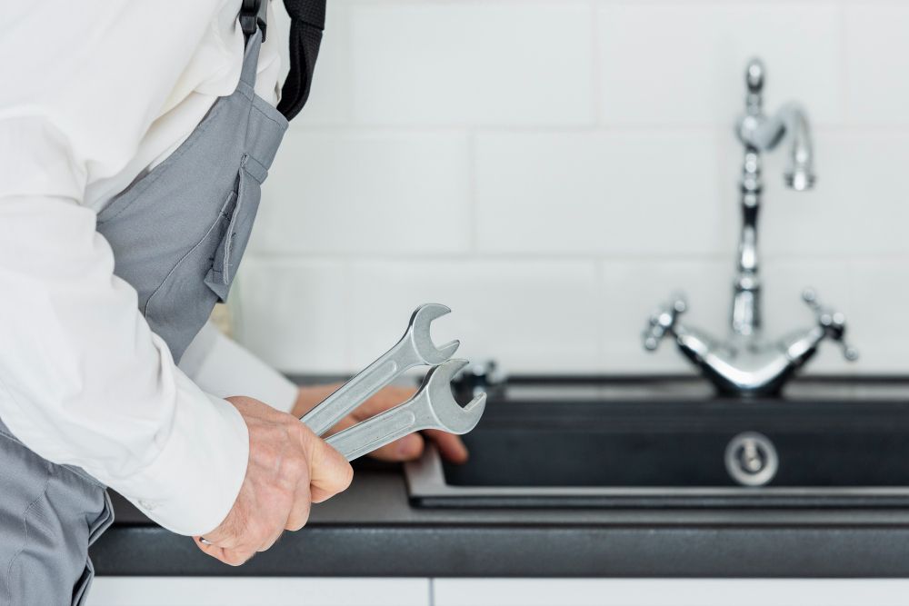 A person in work uniform holding two wrenches next to a kitchen sink with a faucet.
