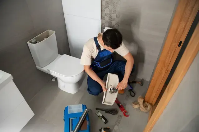 A plumber in blue overalls kneels on a bathroom floor, using a tool to repair a detached toilet tank lid.