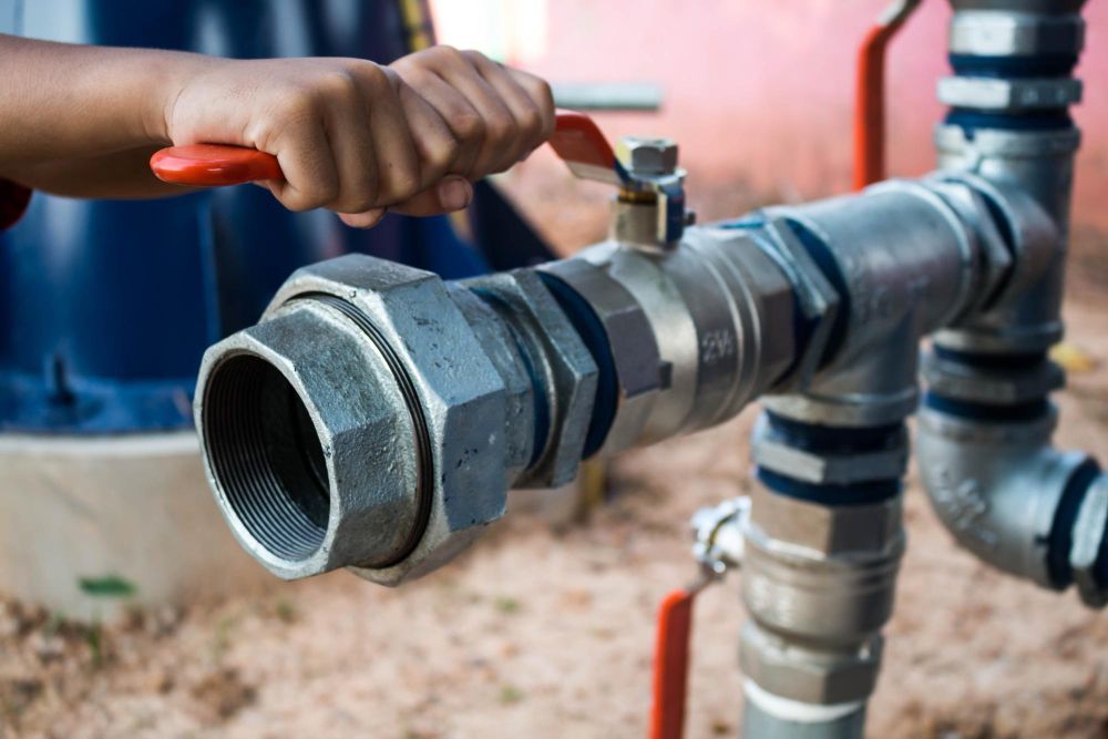 A hand operates the red lever of a silver metal pipe fitting outdoors, showing a plumbing or industrial setup.