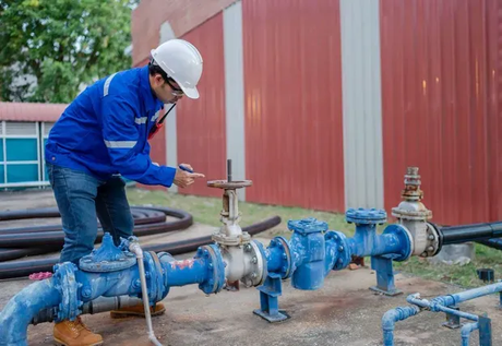 A worker in a blue uniform and white hard hat operates a valve on a blue industrial pipe system outdoors.