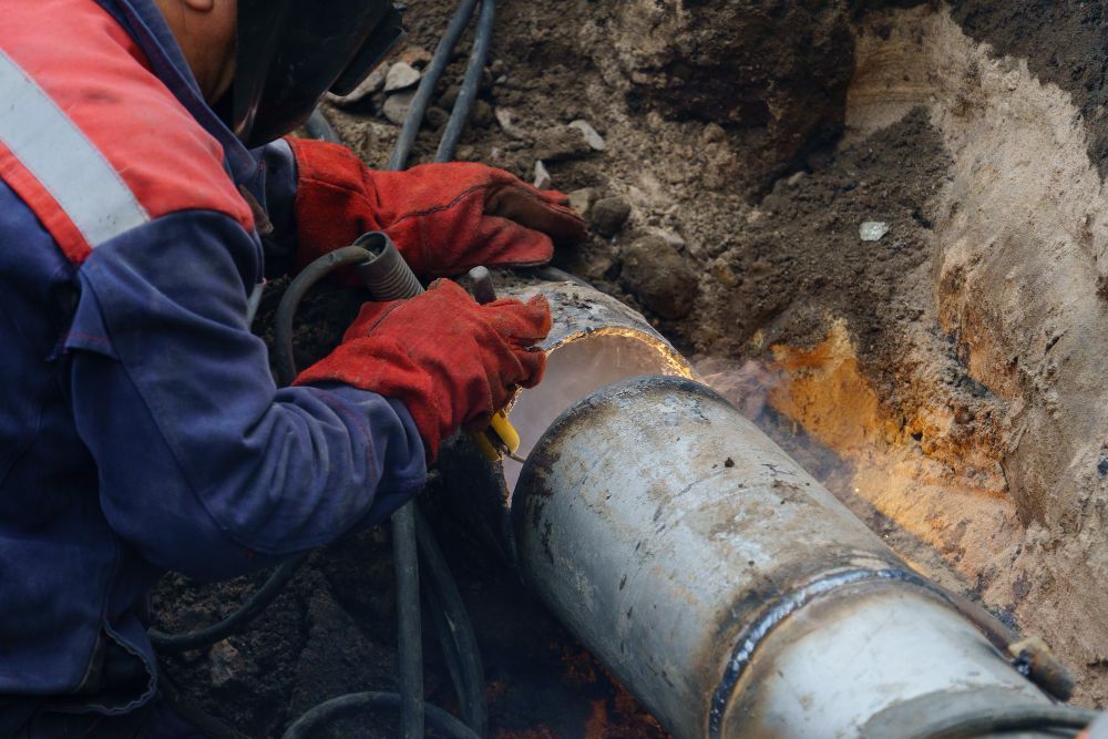 A welder in protective gear and red gloves joins a metal pipe section inside a trench.